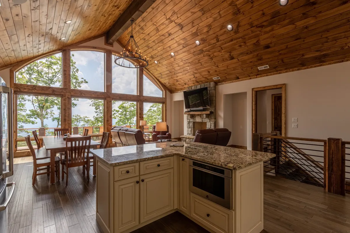 A wide-angle view of the open kitchen, dining, and living area, featuring a central island with granite countertops, a large wooden dining table, comfortable seating, and expansive windows with scenic views. The atmosphere is both inviting and functional, perfect for group activities and celebrations.