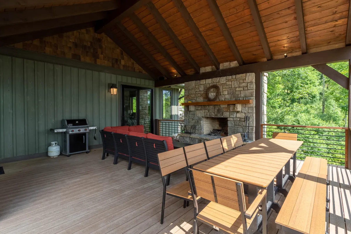 This image captures an outdoor seating area with a grill, stone fireplace, wooden beams, and plush furnishings, surrounded by greenery. The space is designed for al fresco dining, barbecues, and cozy gatherings around the fire.