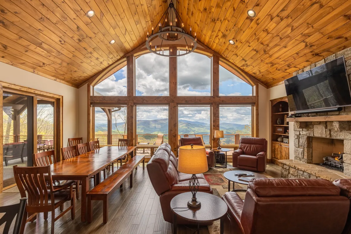 A spacious great room featuring large windows that offer breathtaking mountain views, a rustic wooden dining table that seats a group, comfortable leather seating, and a dramatic stone fireplace. The room is filled with natural light and exudes warmth, making it ideal for family gatherings, dinner parties, and cozy evenings by the fire.