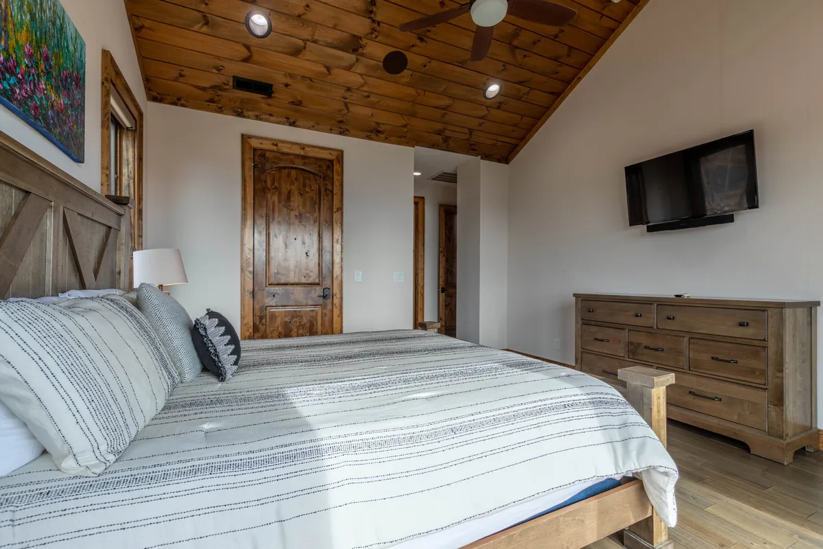 A luxurious bedroom featuring a king-sized bed, warm wooden ceiling, dresser, and a lamp, with a TV positioned for easy viewing. Soft natural light streams through the window, contributing to a serene and comfortable atmosphere, ideal for relaxing or unwinding after a day of activities.