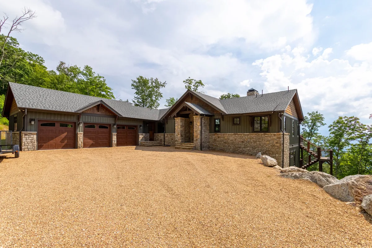 A welcoming exterior view of Clouds Rest featuring a wide stone entryway, wooden garage doors, and a landscaped gravel driveway surrounded by trees. The flat, generous driveway hints at accessible, level parking and easy home entry.