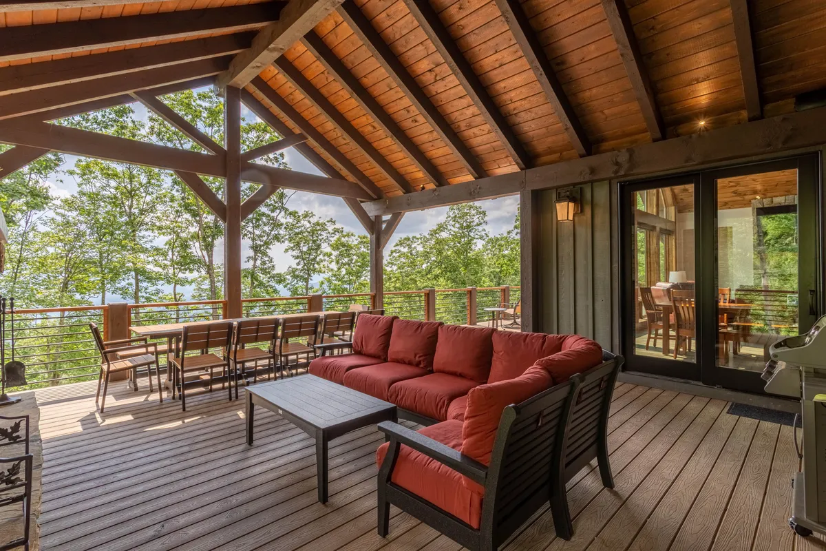 This exterior image showcases a covered deck with wooden beams, a red sofa, a dining table, and lush surrounding trees. The scene is set up for outdoor dining and socializing, with a welcoming and comfortable atmosphere that invites guests to relax, host barbecues, or enjoy an al fresco meal while taking in the natural beauty.