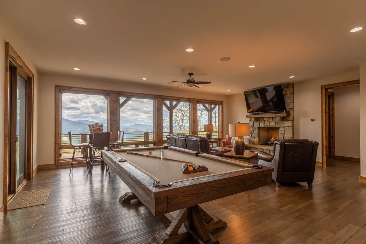 This image showcases a spacious entertainment area featuring a pool table in the foreground, comfortable seating, a glowing fireplace, and large windows that let in natural light. The room has a relaxed and inviting atmosphere—ideal for family gatherings, game nights, and socializing with friends.