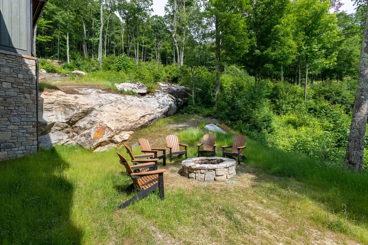 An inviting fire pit area surrounded by wooden chairs and natural rock formations, with lush trees and greenery in the background. The scene exudes a sense of togetherness, relaxation, and outdoor enjoyment, perfect for storytelling or stargazing under the open sky.