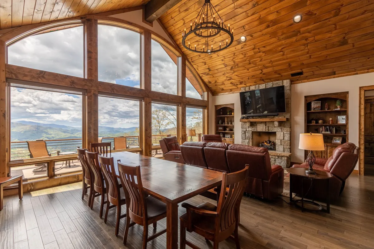 An inviting interior scene showing large windows framing mountain vistas, a long dining table suitable for large groups, plush leather sofas, a fireplace, and a TV. The cozy setting is designed for both celebratory dinners and relaxing movie nights, with abundant space for everyone to gather.