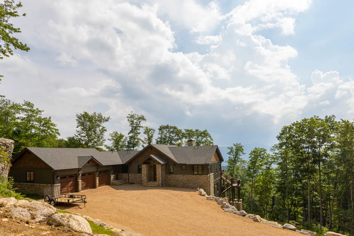 This image showcases the exterior of the house nestled among trees, with a spacious driveway and a backdrop of mountains. The setting highlights open outdoor areas perfect for walking pets and relaxing in nature.