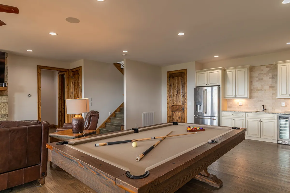 A lively lower-level recreation room with a prominent pool table, leather sofa, adjacent kitchen area, and warm lighting. The space feels casual and fun, ideal for both competition and relaxation, with the kitchen amenities visible for easy access to snacks and drinks.